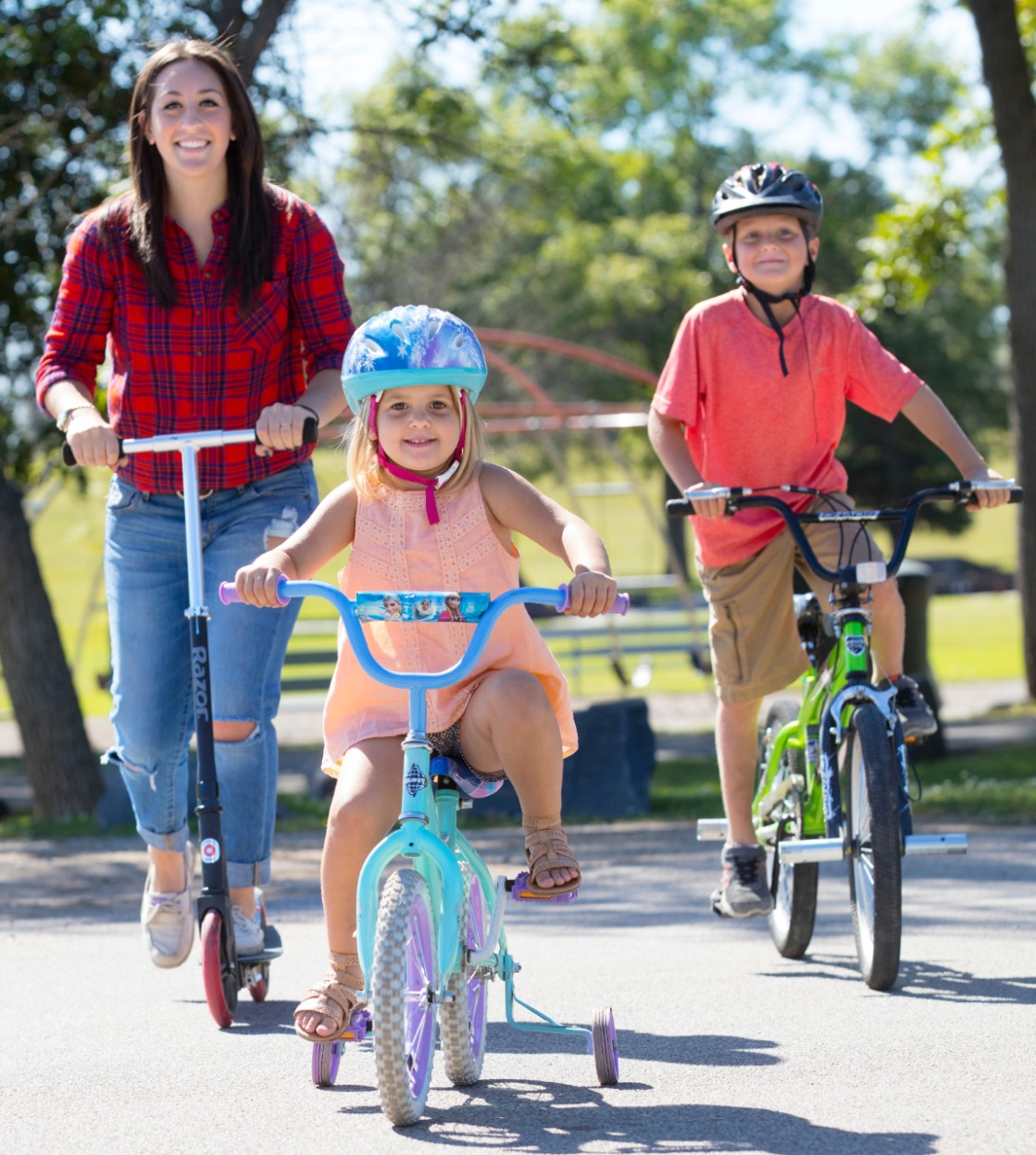 Two young kids riding scooters and bikes with an adult riding bike in the park on a sunny day